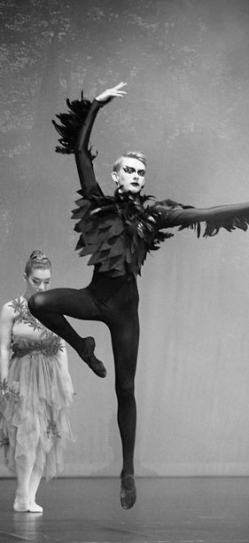 Ballettänzer in dunklem Kostüm springt, ein Bein angewinkelt, ein Arm zur Seite, der andere nach oben.